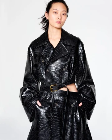 An Asian woman models a double-breasted, black, crocodile-texture trench coat with a belt and matching skirt against a white backdrop.