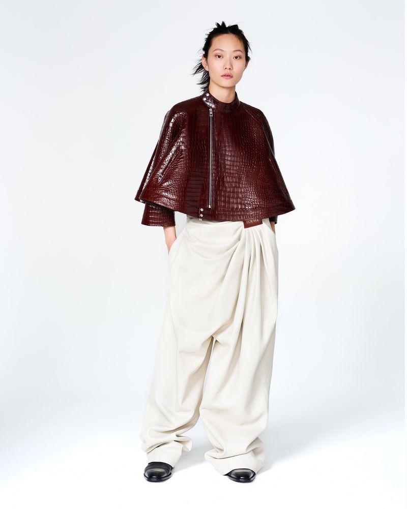 Asian model poses in a studio wearing a red-brown croc leather cape, cream-colored draped pants, and black flats against a white backdrop.