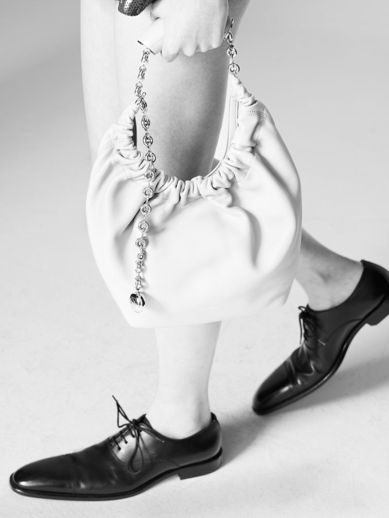 Model holding a slouchy white leather shoulder bag with a chain strap, wearing black leather oxford shoes against a plain background.