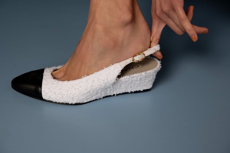 Close-up of a woman's foot partially inside a slingback wedge heel shoe with a black cap toe and white textured body on a grey background.
