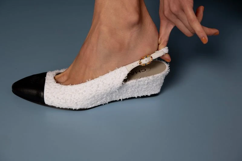 Close-up of a woman's foot partially inside a slingback wedge heel shoe with a black cap toe and white textured body on a grey background.