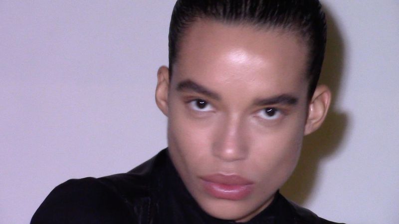 Close-up portrait of a young adult with slicked-back dark hair and smooth skin, wearing black clothing against a pale backdrop.