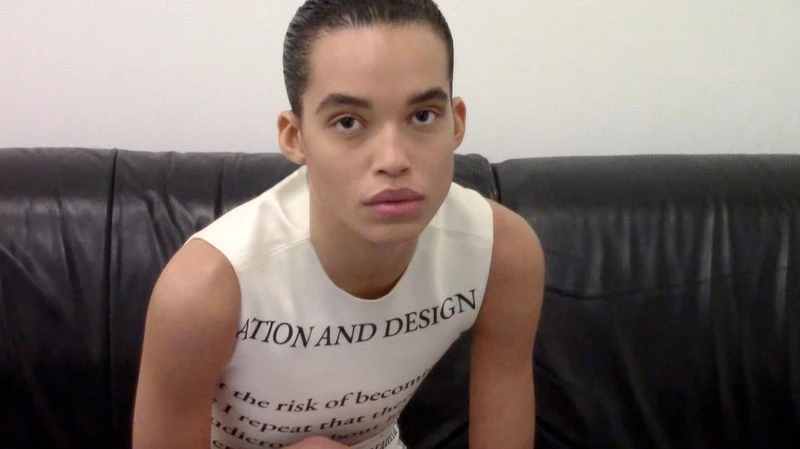 Close-up shot of an adult male with slicked-back dark hair, wearing a white sleeveless shirt with black text while sitting on a black couch.