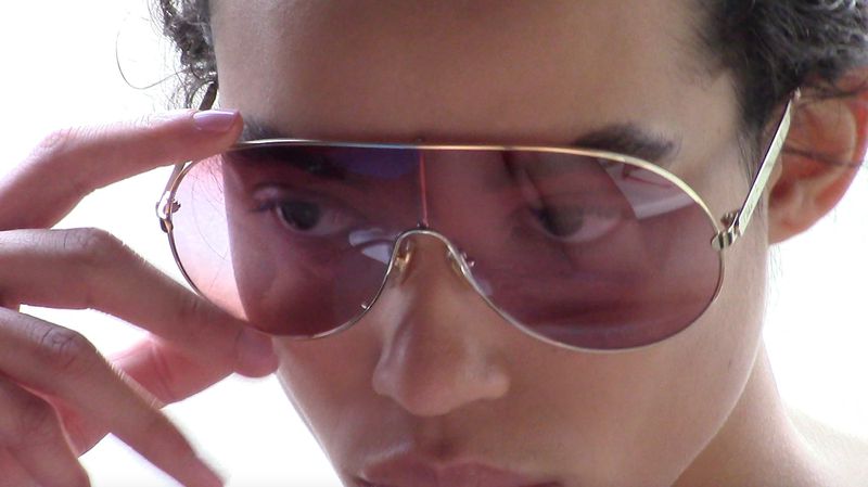 Close-up of a woman wearing aviator sunglasses with a gold frame and pink lenses, her hand gently touching the frame.