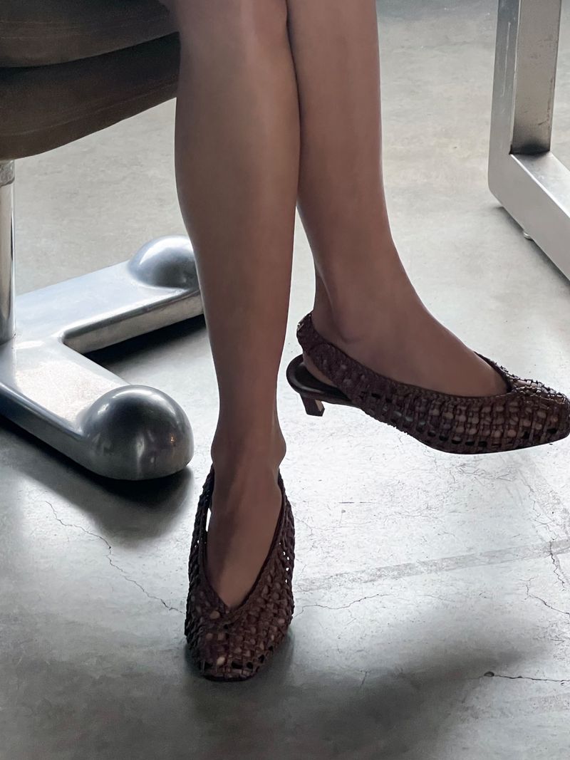 Close-up of woman's legs and feet wearing brown woven slingback shoes while seated on a metal-legged chair on a grey floor.
