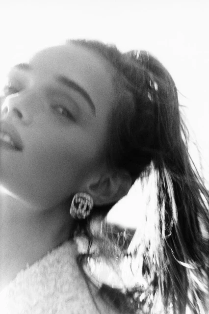 Black and white portrait of a young woman with dark hair, wearing statement earrings, looking up towards a bright, sunny sky.