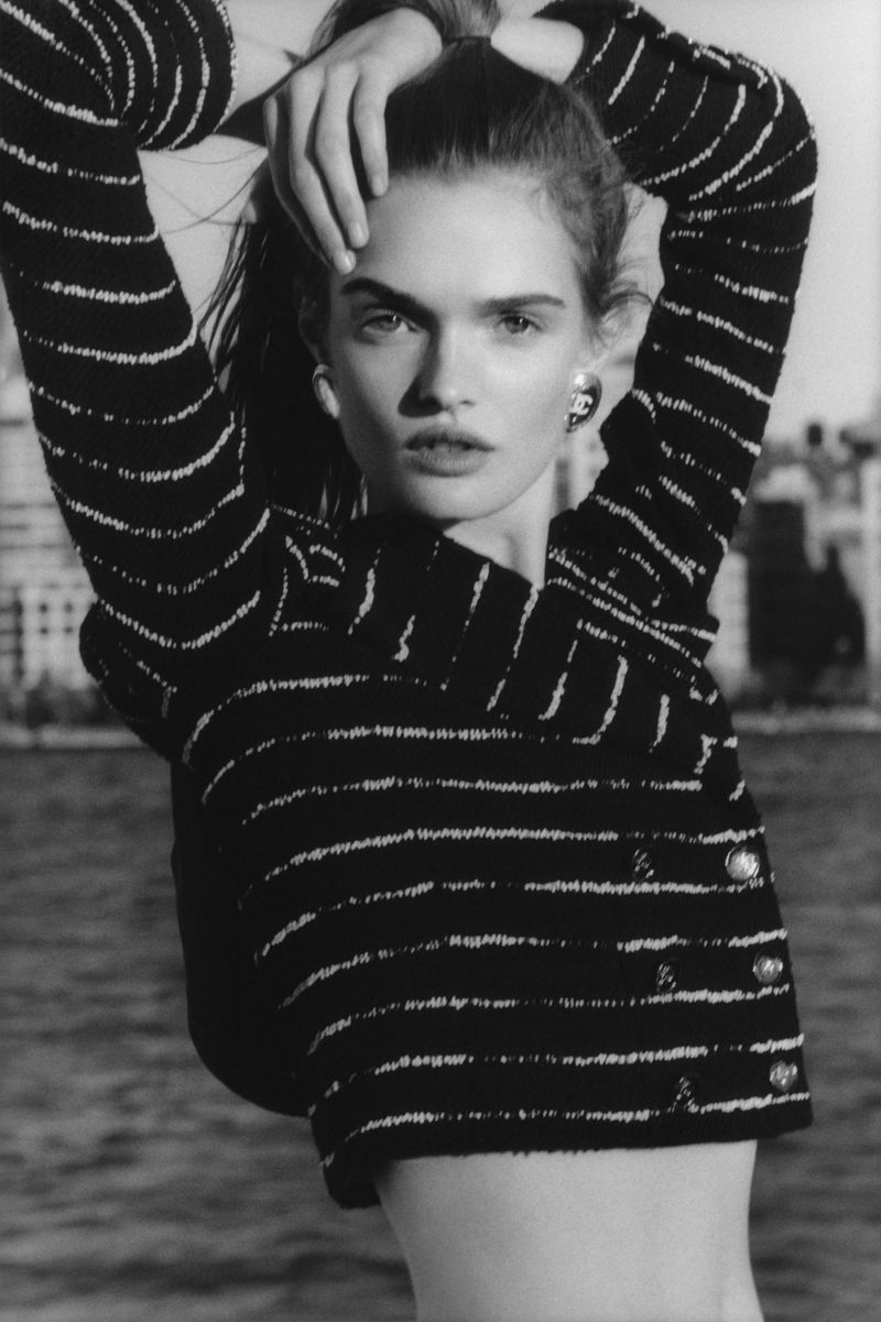 Monochrome portrait of a woman with a striped jacket, posing against an outdoor background of water and buildings, captured in high contrast.