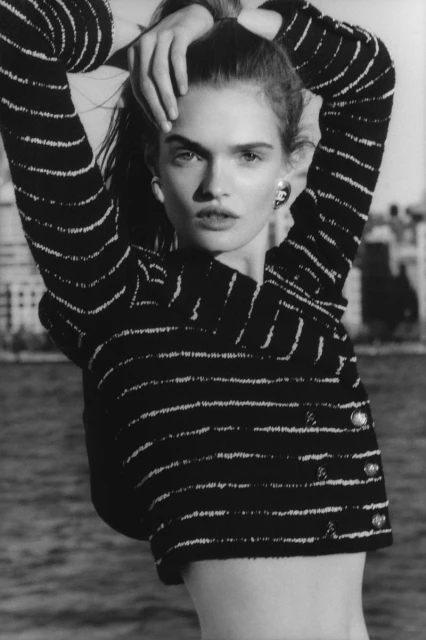 Monochrome portrait of a woman with a striped jacket, posing against an outdoor background of water and buildings, captured in high contrast.