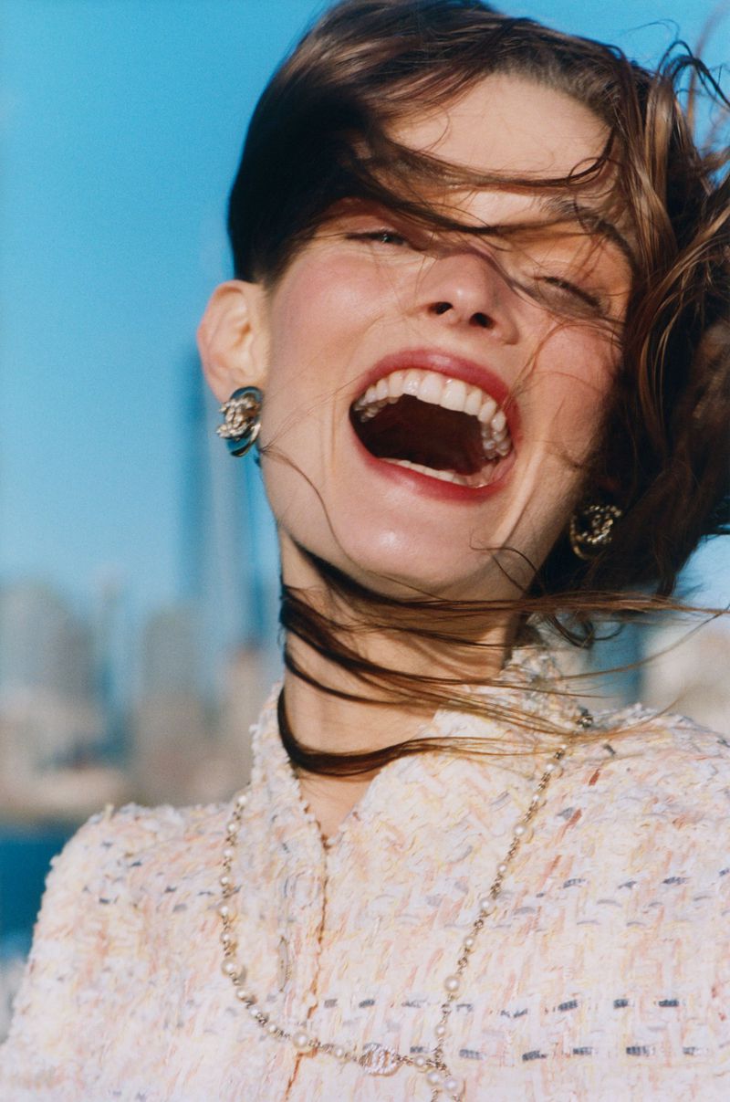 Smiling woman with windblown hair wears a cream-colored Chanel jacket, statement earrings, and necklace against a blue sky and cityscape.