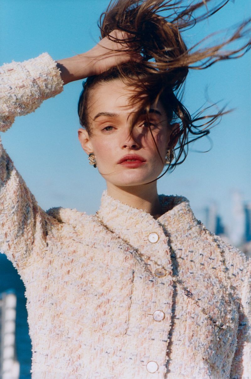 Headshot of a woman with hair up, wearing a tweed jacket with pearl buttons against a blue sky background, in a fashion editorial style.