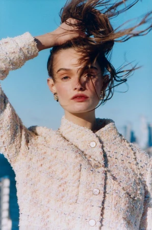 Headshot of a woman with hair up, wearing a tweed jacket with pearl buttons against a blue sky background, in a fashion editorial style.