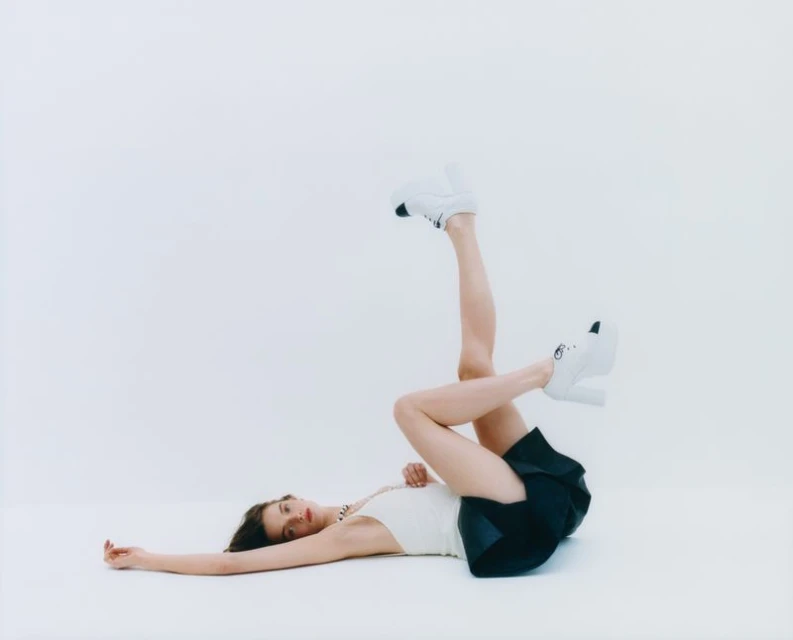 Woman in white top and black shorts poses barefoot on white backdrop, legs raised with platform shoes, relaxed arm.