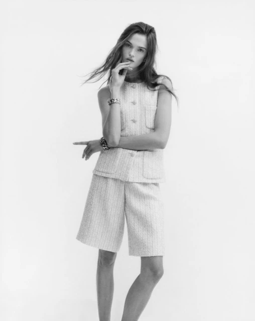 Monochrome portrait of an adult female fashion model wearing a tweed vest and shorts, posing in a photo studio against a white backdrop.
