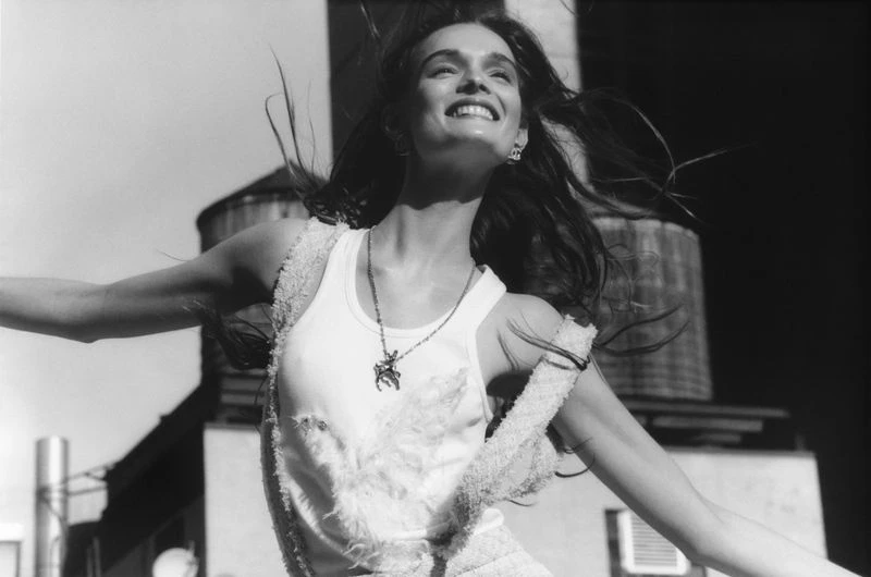 A woman smiles with arms out on a rooftop with a water tower in the background, monochrome, joyful and optimistic. 