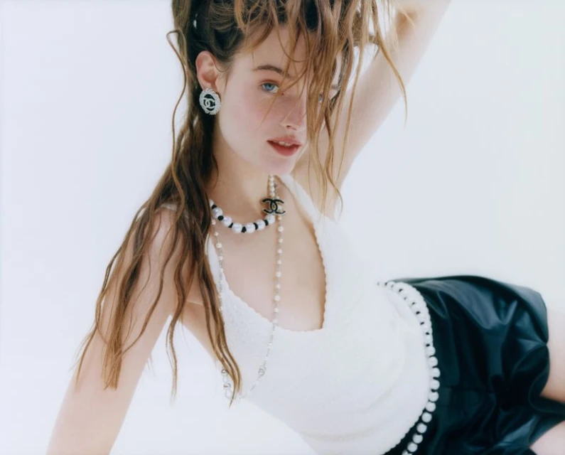 Close-up studio shot of a young adult female model with wet hair, white tank top, black skirt, and Chanel jewellery on a white background.