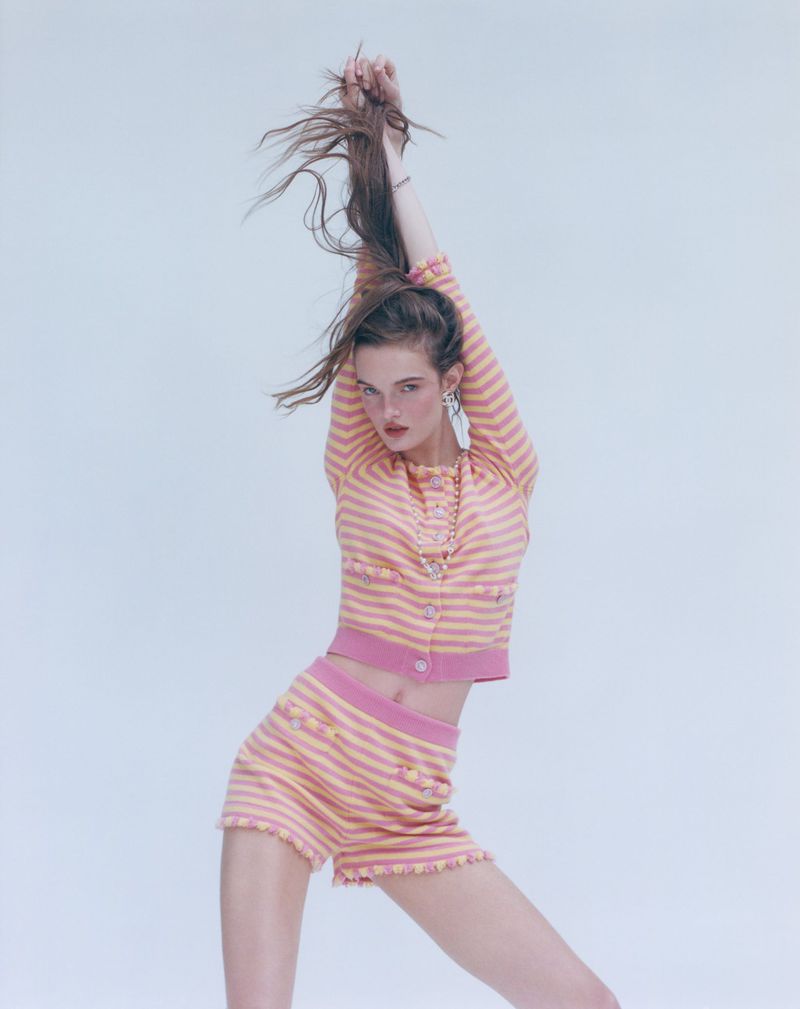 A slender woman poses in a pink and yellow striped cardigan and shorts set against a light backdrop, her arms raised above her head.
