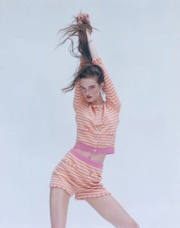 A slender woman poses in a pink and yellow striped cardigan and shorts set against a light backdrop, her arms raised above her head.