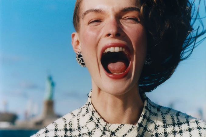 Close-up portrait of model Lulu Tenney laughing with mouth open against a blue sky with the Statue of Liberty visible in the background.