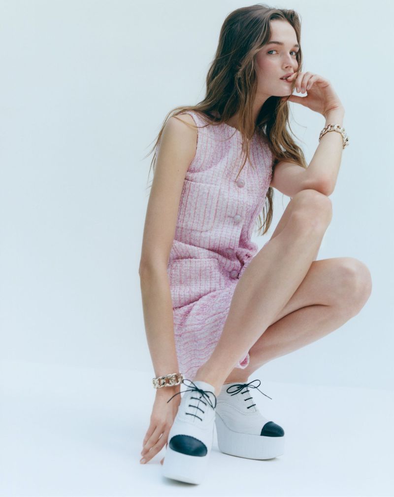 A young woman with long wavy hair wears a pink tweed dress and white platform shoes in a bright studio setting, fashion shot.