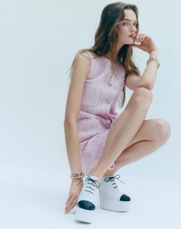 A young woman with long wavy hair wears a pink tweed dress and white platform shoes in a bright studio setting, fashion shot.
