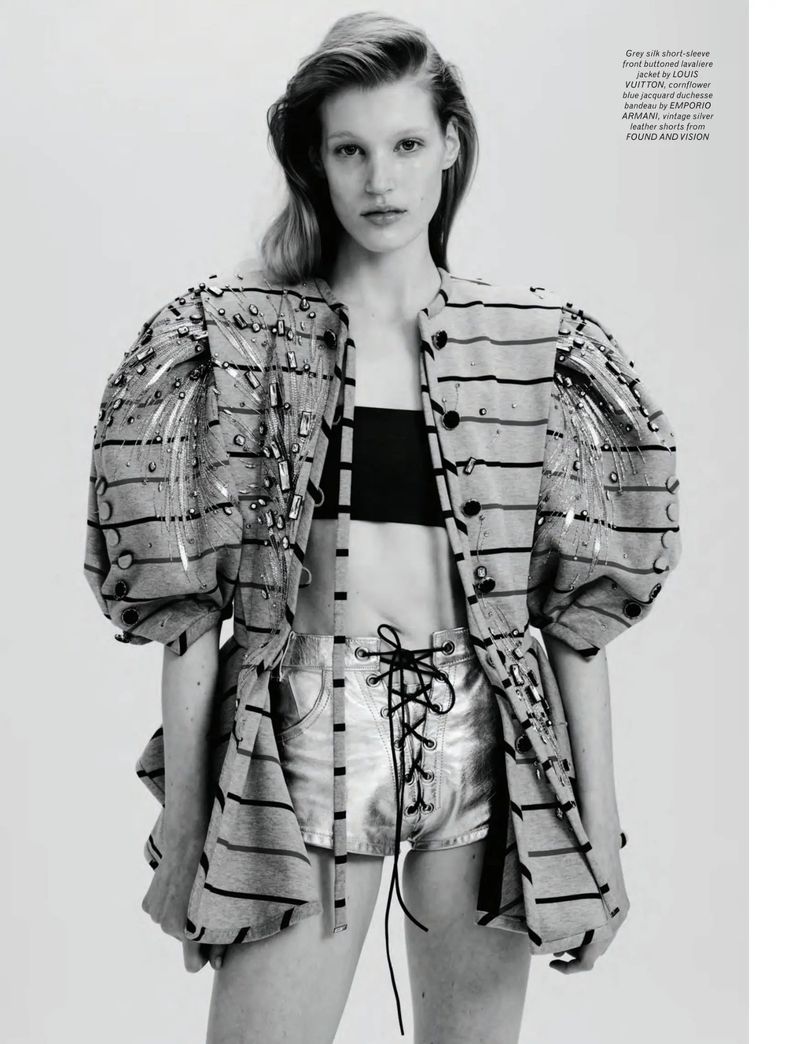 Fashion editorial shot of a blonde model in silver shorts and a striped jacket posing in a photo studio with an off-white background.