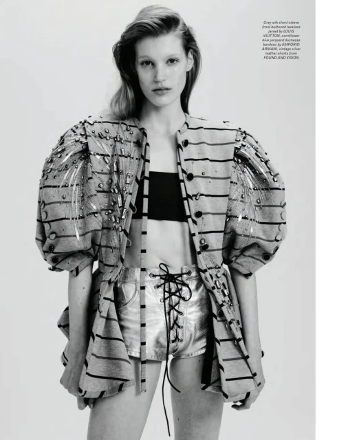 Fashion editorial shot of a blonde model in silver shorts and a striped jacket posing in a photo studio with an off-white background.
