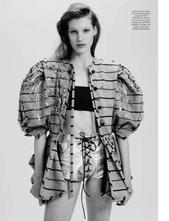 Fashion editorial shot of a blonde model in silver shorts and a striped jacket posing in a photo studio with an off-white background.