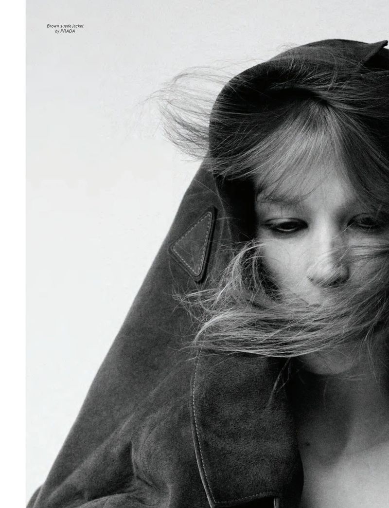 Close-up monochrome portrait of model Jacqui Hooper with hair blowing across her face wearing a brown jacket against a white background