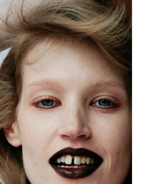 Close-up portrait of a blonde woman with light skin and gap teeth, wearing dark lipstick. Beauty shot with a bright and neutral background.