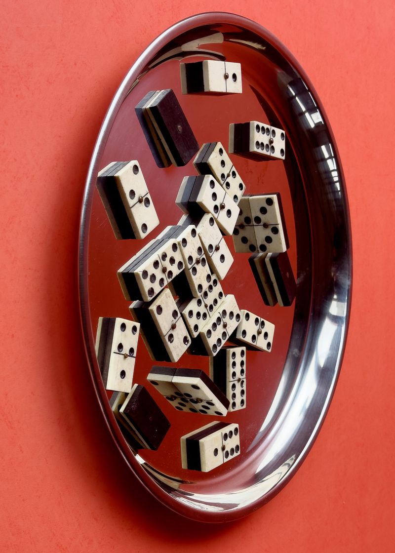 Dominoes arranged on a shiny silver tray against a vivid orange background, creating a still life composition with geometric shapes.