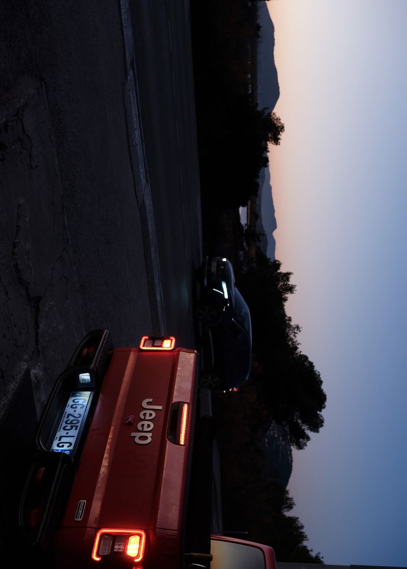 Red Jeep pickup truck parked on a road at dusk, with a sedan and mountain range in the background. Sky features gradient from yellow to blue.