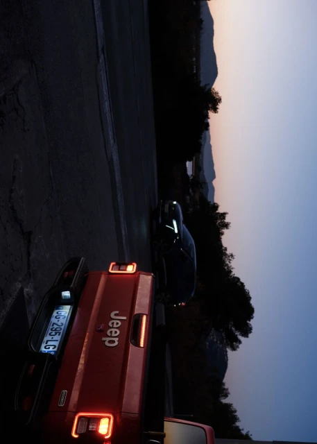 Red Jeep pickup truck parked on a road at dusk, with a sedan and mountain range in the background. Sky features gradient from yellow to blue.