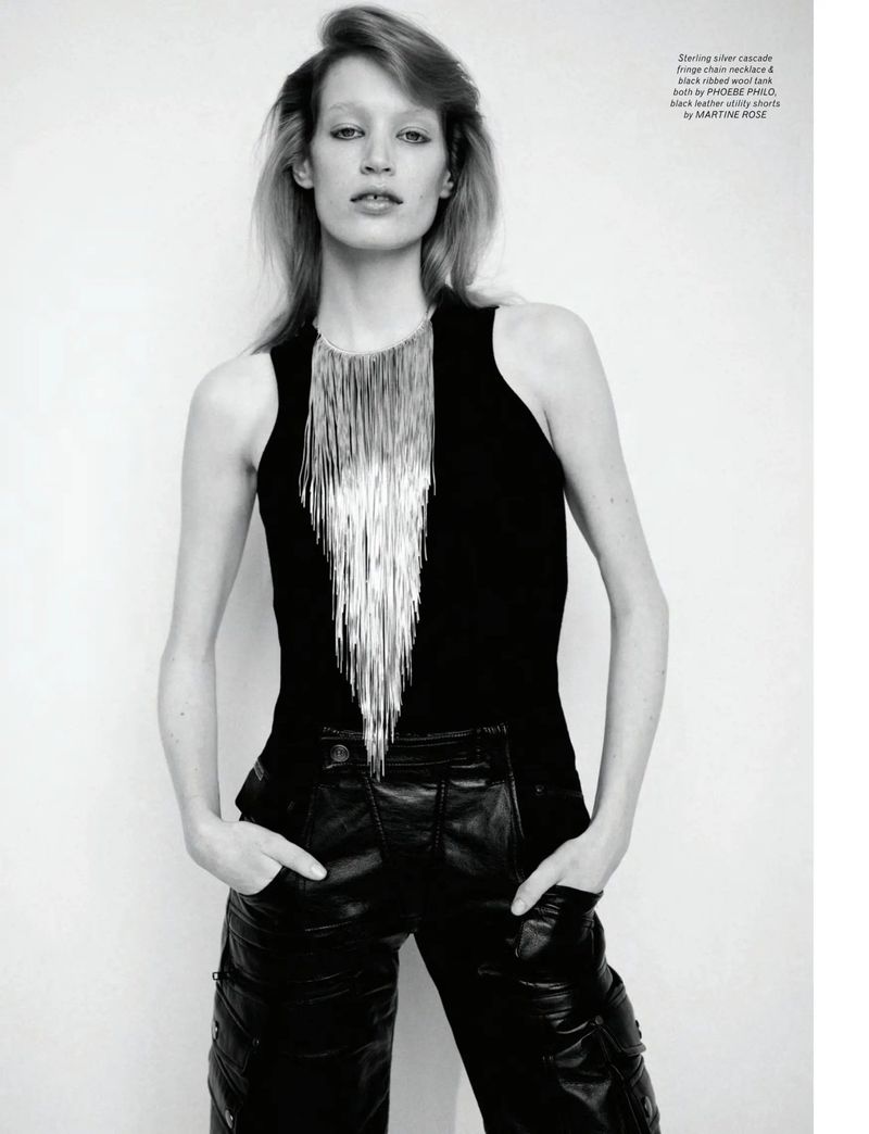 Fashion shot of a slender woman with blonde hair in a black tank top, silver fringe necklace, and black leather pants against a white wall.