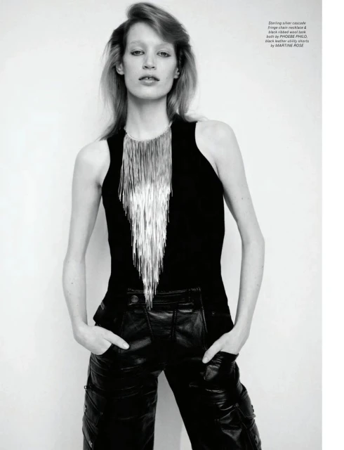 Fashion shot of a slender woman with blonde hair in a black tank top, silver fringe necklace, and black leather pants against a white wall.