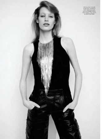 Fashion shot of a slender woman with blonde hair in a black tank top, silver fringe necklace, and black leather pants against a white wall.