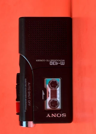 Vintage black Sony M-430 microcassette recorder with a small cassette visible, set against a solid orange background in a studio shot.
