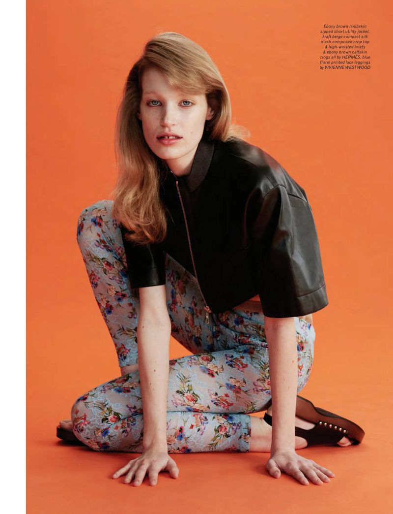 Model Jacqui crouched against a solid orange background wearing a black leather jacket and floral print leggings for Pop Magazine.