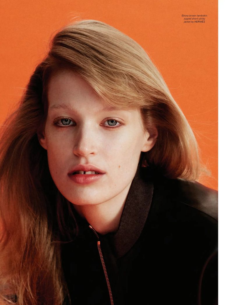 Close-up portrait of a fair-skinned, light-haired woman in a dark bomber jacket against an orange background in a fashion editorial.