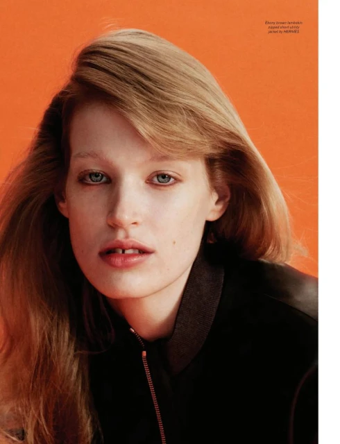 Close-up portrait of a fair-skinned, light-haired woman in a dark bomber jacket against an orange background in a fashion editorial.