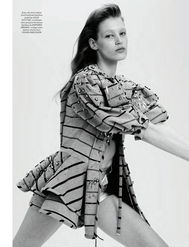 Fashion editorial shot of a woman in a striped jacket and shiny shorts, striking a dynamic pose in a brightly lit photo studio.