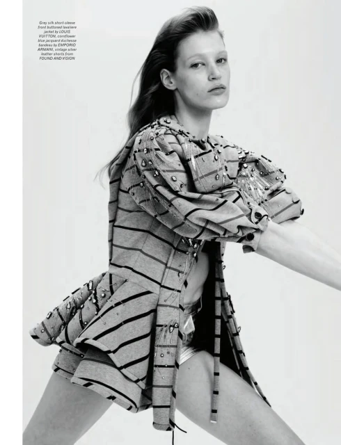 Fashion editorial shot of a woman in a striped jacket and shiny shorts, striking a dynamic pose in a brightly lit photo studio.