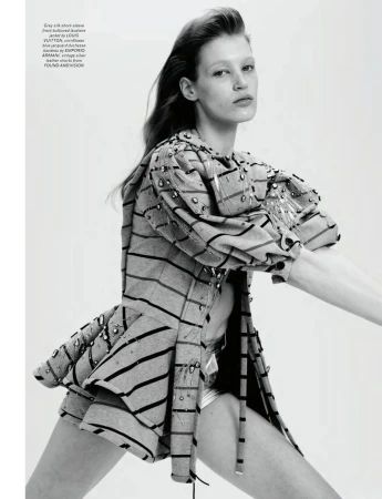 Fashion editorial shot of a woman in a striped jacket and shiny shorts, striking a dynamic pose in a brightly lit photo studio.