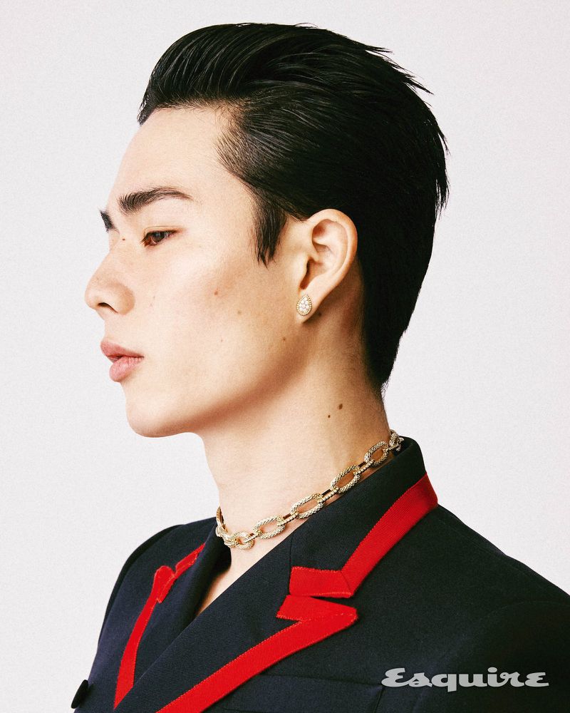 Profile shot of a young Asian man with slicked-back hair wearing a black jacket with red trim, a gold chain, and an earring against white.