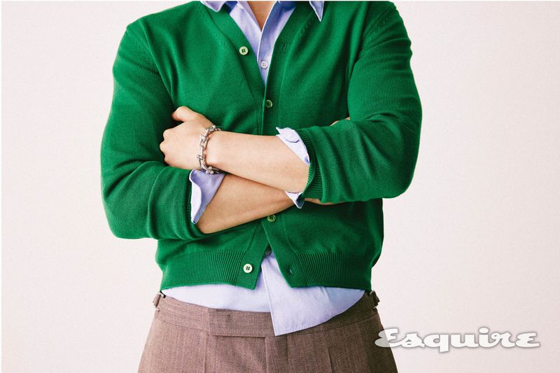 Man in a green cardigan and brown slacks with arms crossed in a studio shot. The Esquire logo is visible in the lower right corner.