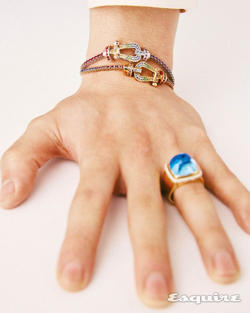 Close-up shot of a person's hand displaying gold bracelets with colorful gem details, along with a gold ring with a blue stone, on a white background.