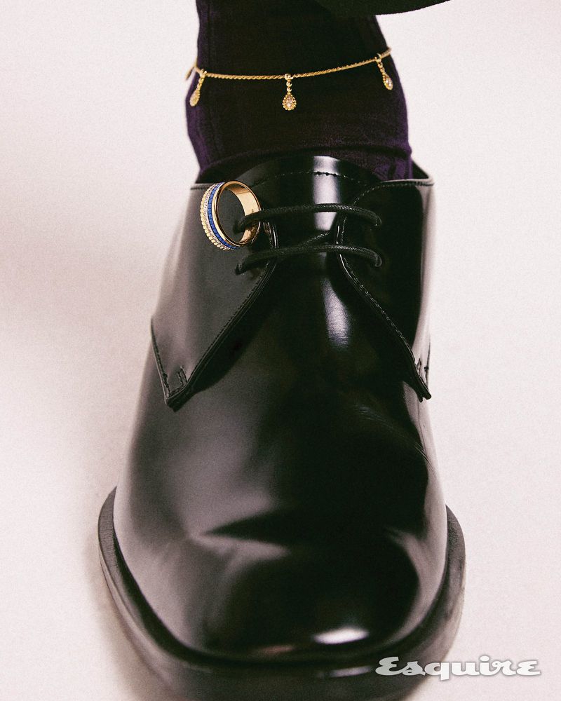 Close-up of a polished black leather shoe with a gold and blue ring on the laces, paired with a black sock and delicate gold chain.