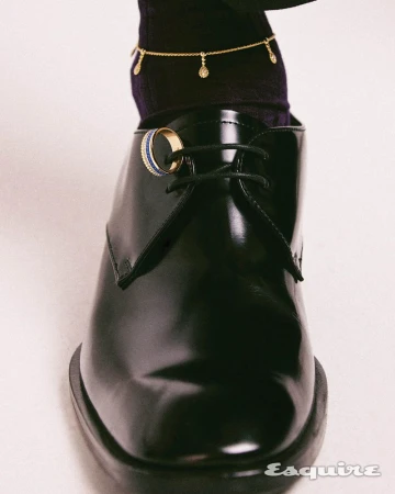 Close-up of a polished black leather shoe with a gold and blue ring on the laces, paired with a black sock and delicate gold chain.