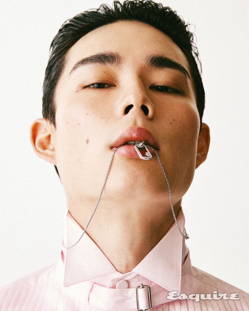 Close-up portrait of an Asian man with short hair, wearing a pink shirt and silver chain necklace, against a plain white backdrop.