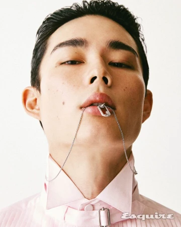 Close-up portrait of an Asian man with short hair, wearing a pink shirt and silver chain necklace, against a plain white backdrop.
