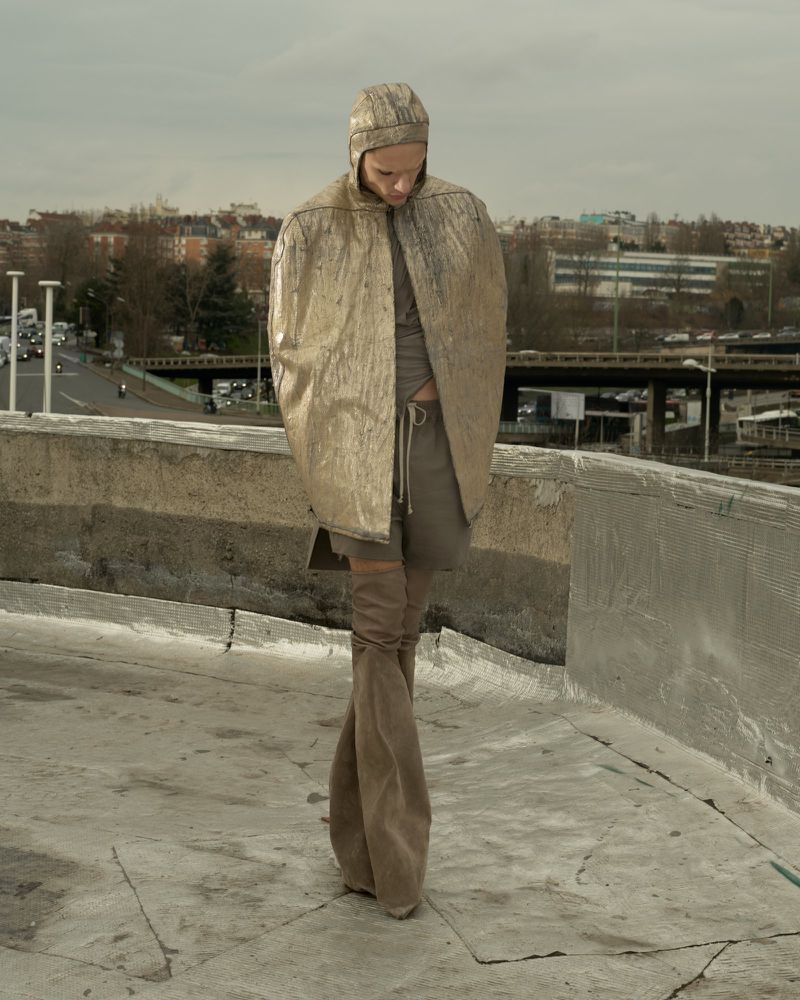 Fashion shot on a rooftop featuring a woman wearing a hooded gold cape and thigh-high boots against a blurred city backdrop.
